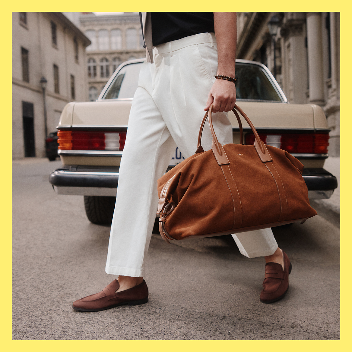 A person walking outdoors carrying a large brown suede duffel bag with tan leather handles, wearing white trousers and brown loafers, with a vintage car in the background.