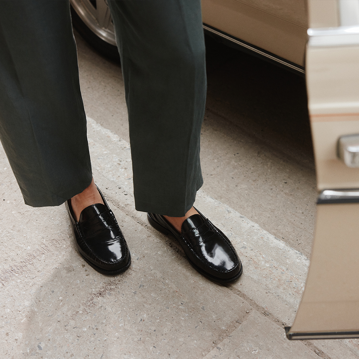 Une personne portant des mocassins en cuir verni noir avec un pantalon gris anthracite foncé, debout sur une surface en béton à côté d’un véhicule beige.