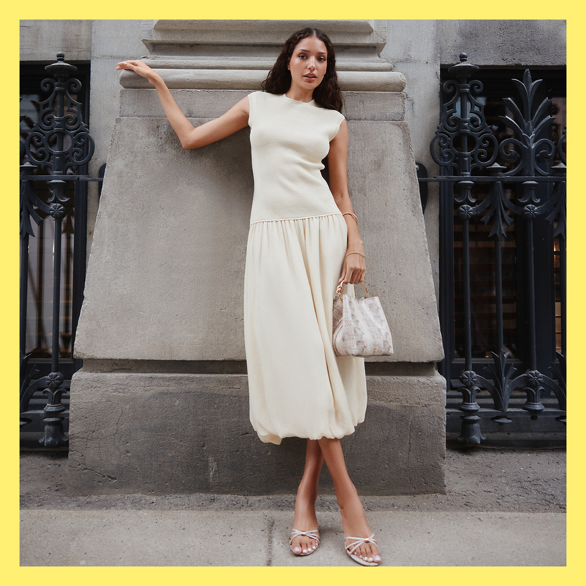 A woman stands outdoors against a stone building and wrought iron gate, wearing a sleeveless cream midi dress with a gathered waist and kitten heels while holding a small handbag.