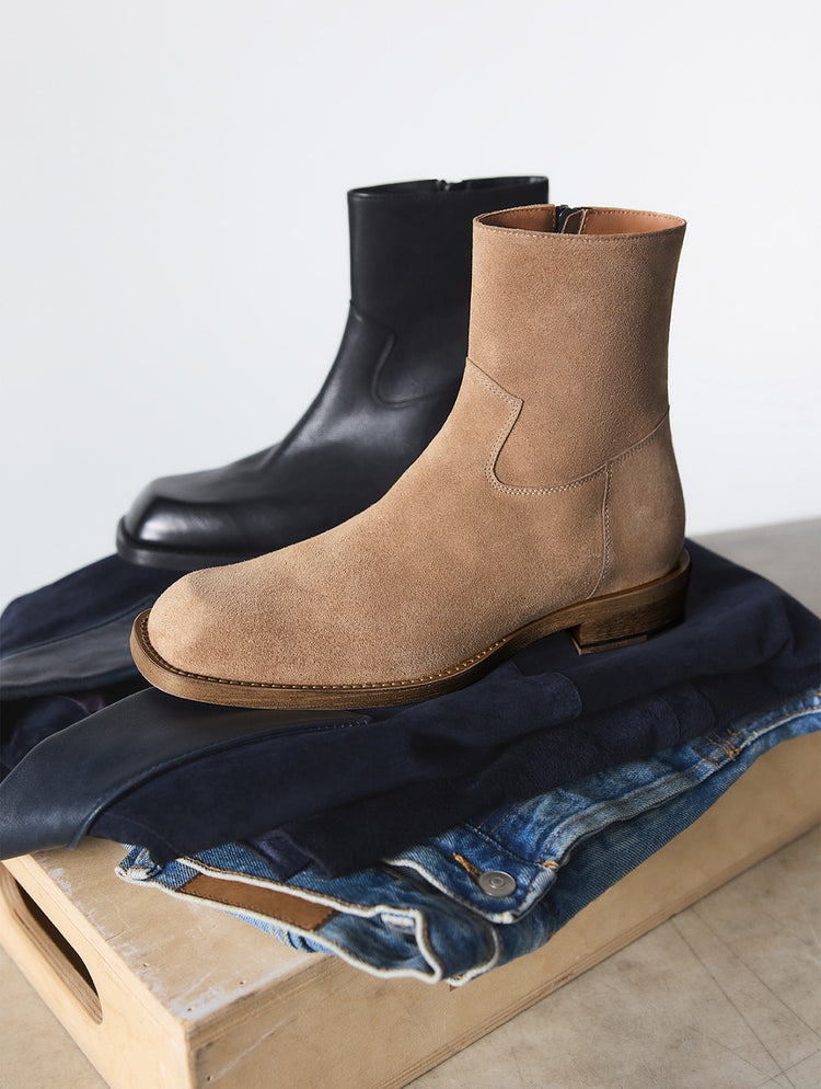 Une paire de bottillons de style Chelsea, l’un en suède tan et l’autre en cuir noir, mise en scène sur un empilement de jeans bleu marine et bleu repliés, déposés sur une caisse en bois pâle devant un fond blanc uni.