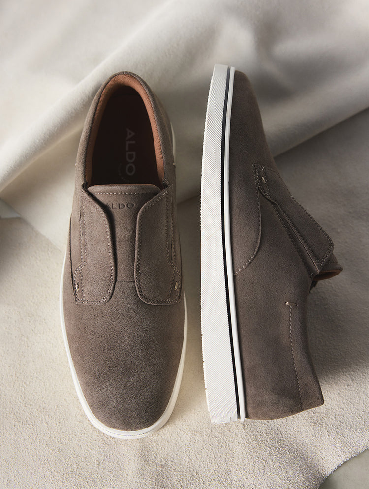 Une paire d’espadrilles à enfiler de la marque ALDO en suède brun taupe, présentée en vue de dessus et de côté sur un tissu beige drapé.