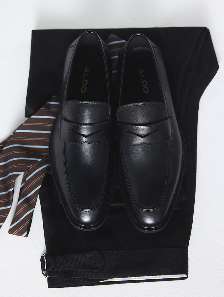 Une paire de mocassins penny en cuir noir de la marque ALDO, présentée sur un veston et un pantalon de complet noirs, accompagnés d’une cravate rayée brun, bleu et blanc. La photo est prise en plongée sur un fond blanc.