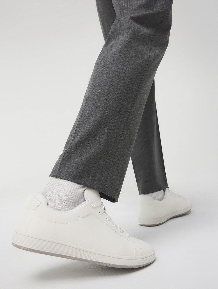 Des espadrilles blanches en cuir à tige basse portées avec des chaussettes blanches mi-mollet et un pantalon gris, photographiées en contre-plongée sur un fond de studio gris pâle.