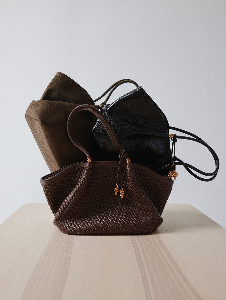 Une pile de trois sacs à main de créateur, comprenant à l’avant un fourre-tout en cuir tressé brun, derrière lui un sac en suède brun foncé, et à l’arrière un sac noir à effet crocodile embossé, présentés sur une surface en bois pâle devant un fond de studio neutre.