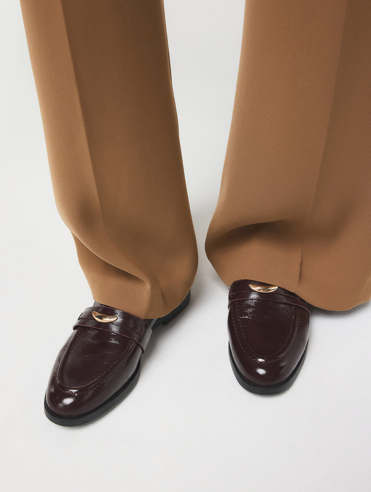 Une vue rapprochée des pieds d’une personne portant des mocassins penny en cuir verni brun foncé ornés de détails métalliques de couleur or, agencés à un pantalon tan à jambe large, sur un fond de studio gris pâle.