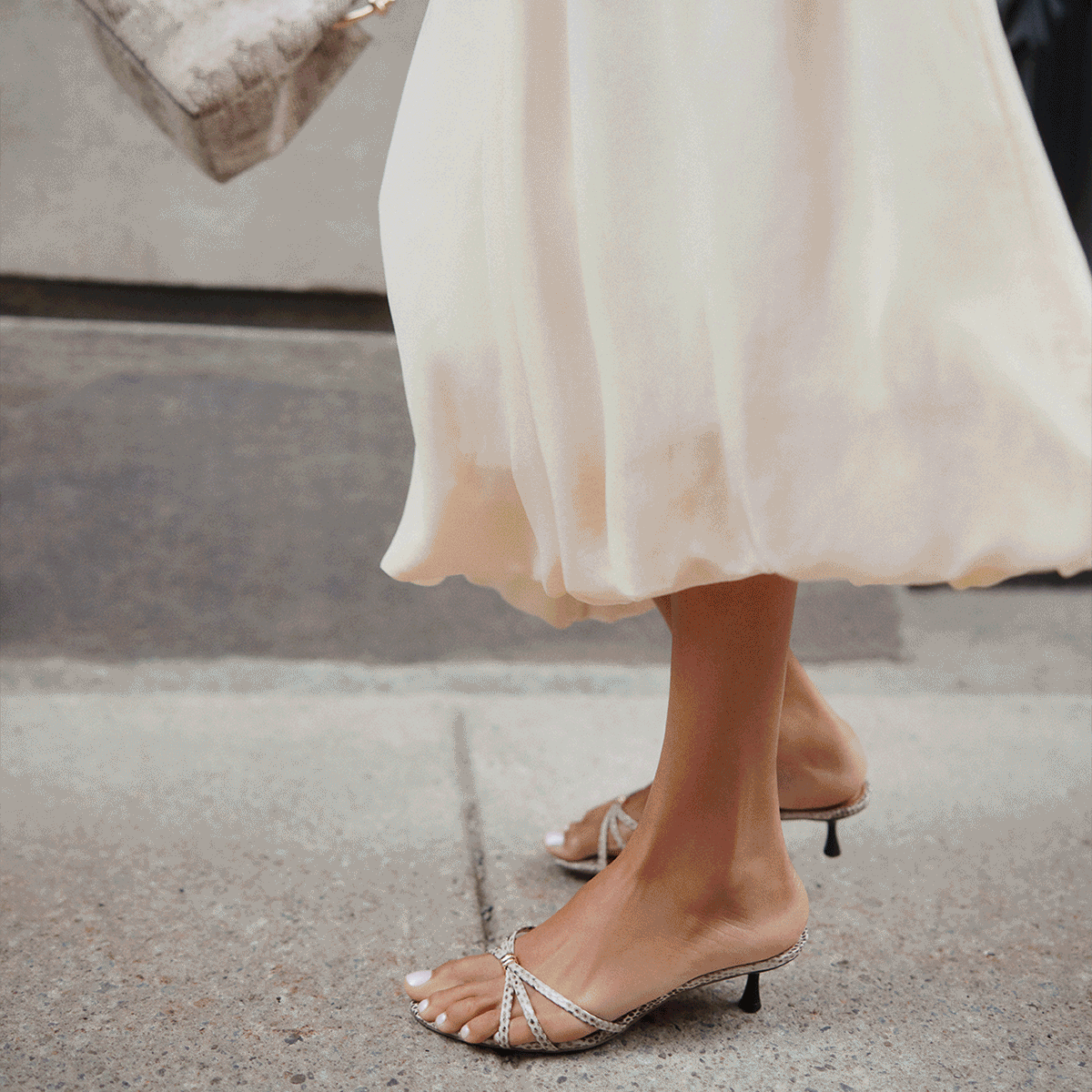 Une image GIF de style de vie montrant une femme portant une robe midi crème sans manches et tenant un sac à main blanc, ainsi qu’un homme en pantalon blanc transportant un sac de voyage en suède brun dans une rue urbaine.