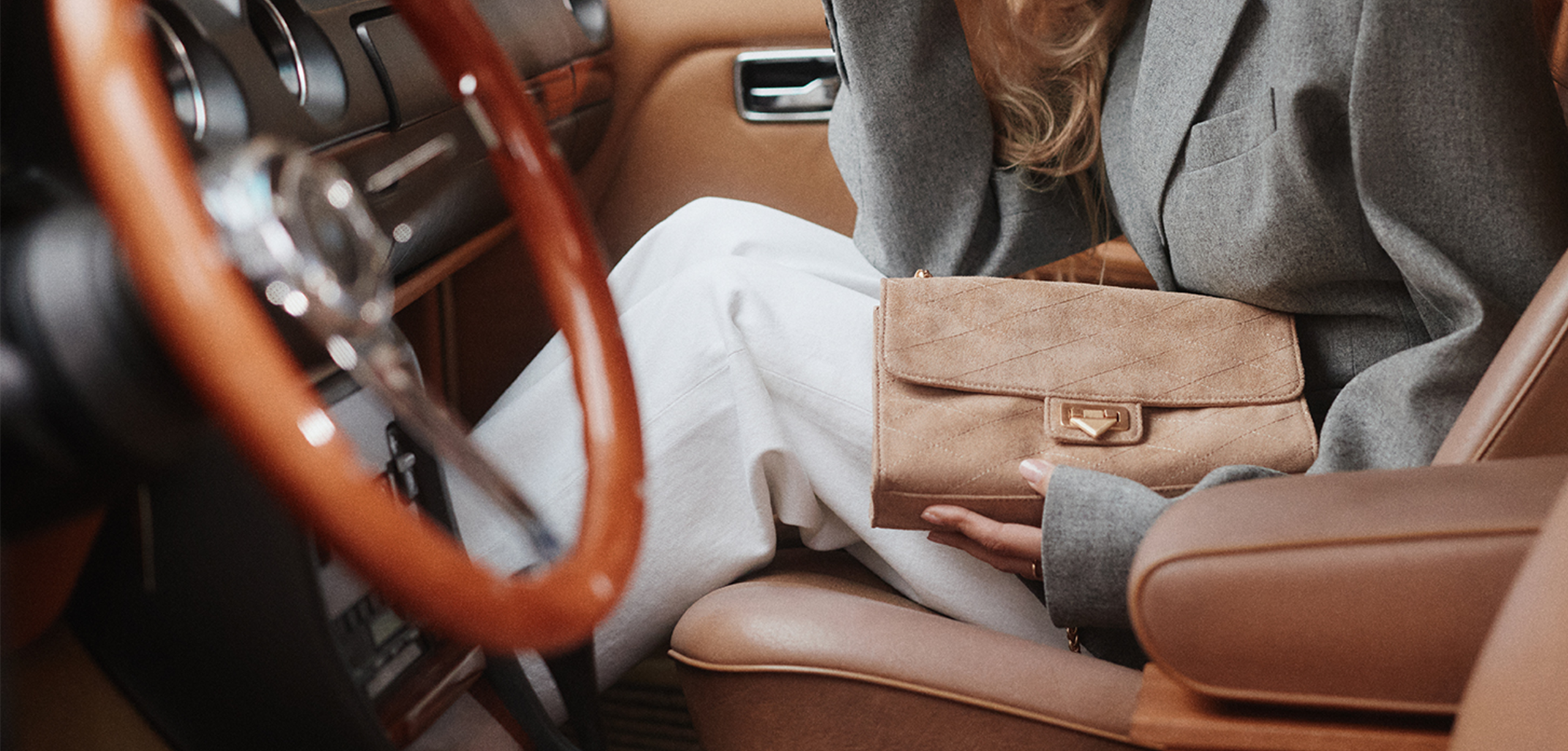 A woman sitting inside a luxury car with brown leather seats holds a small taupe suede purse.