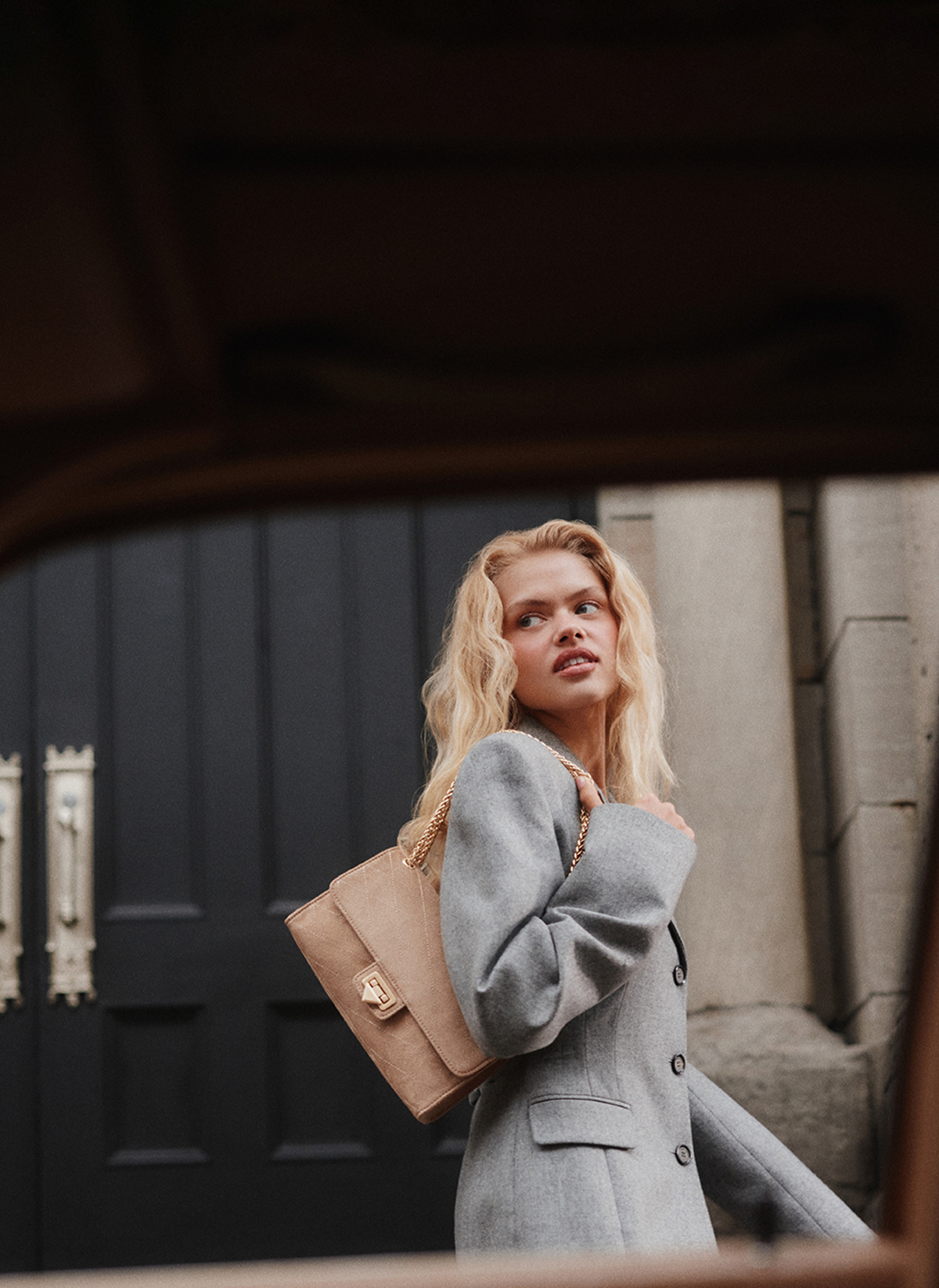 A blonde woman wearing a light gray coat and carrying a beige shoulder bag with a gold clasp, standing in front of a dark paneled door.