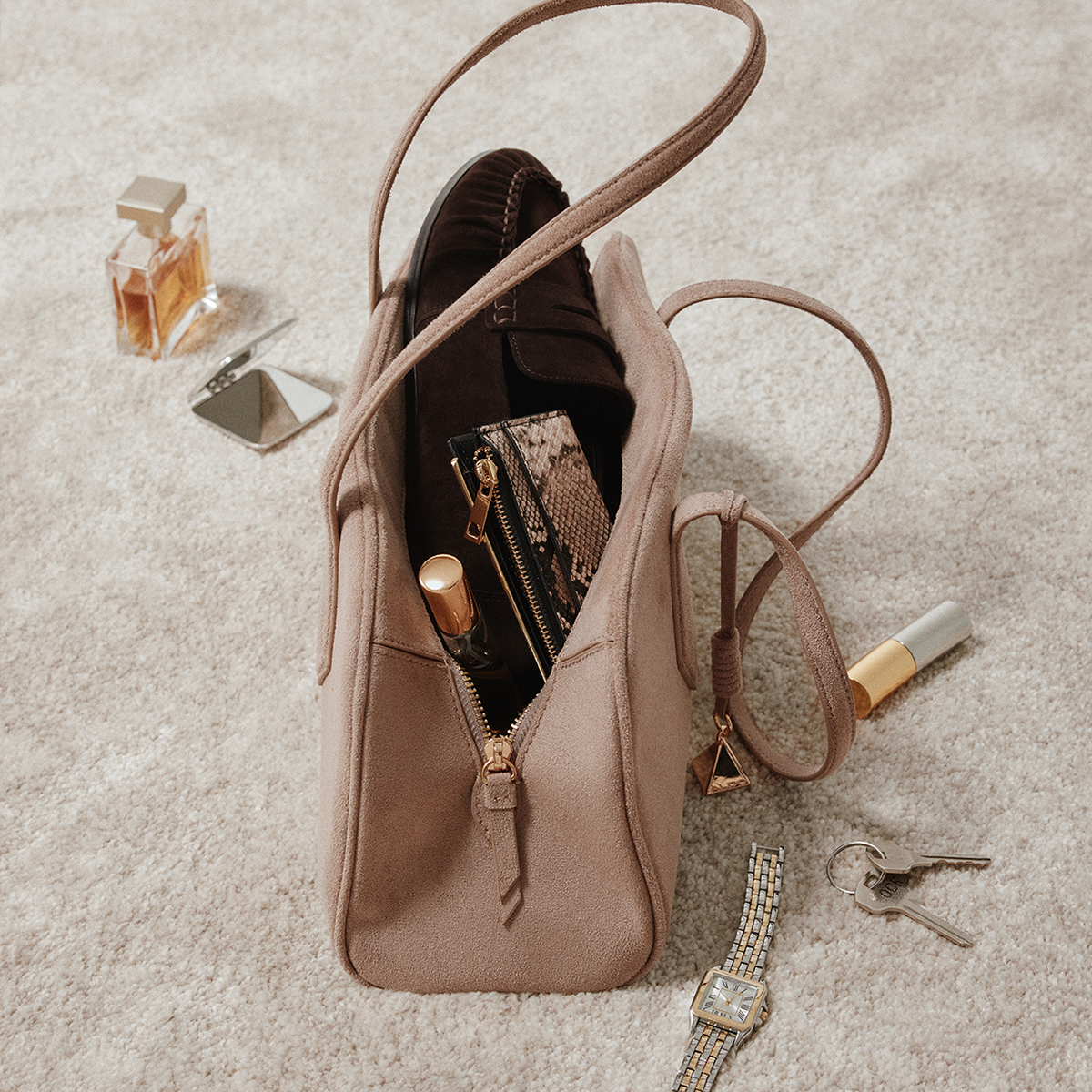 A taupe suede shoulder bag with a zippered main compartment, shown open with various cosmetics and accessories inside. The bag is styled on a textured beige carpet with a perfume bottle, keys, a watch, and a lipstick nearby.