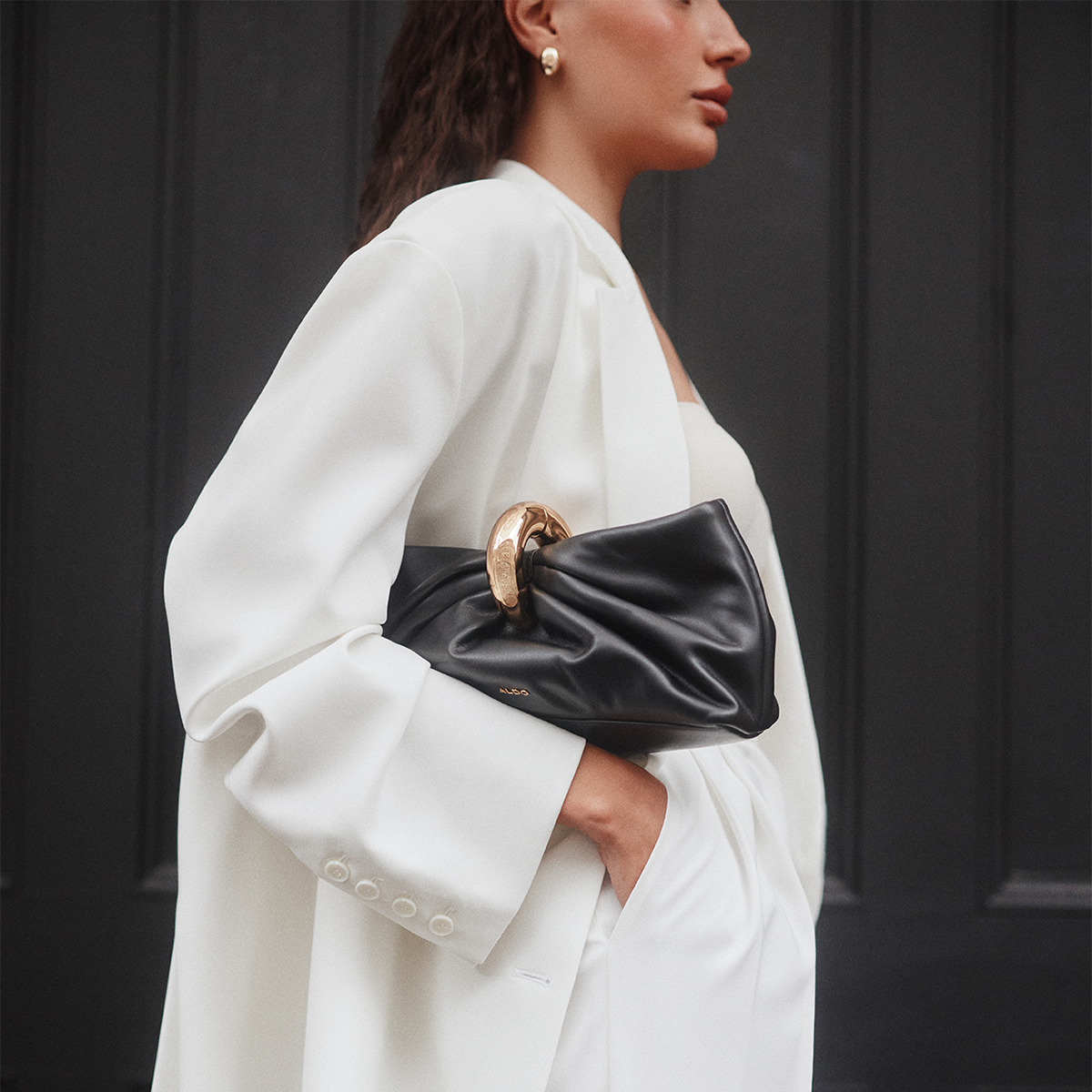 A woman in a white blazer and white dress carries a black leather clutch bag with a prominent gold-tone circular ring handle against a dark paneled door.