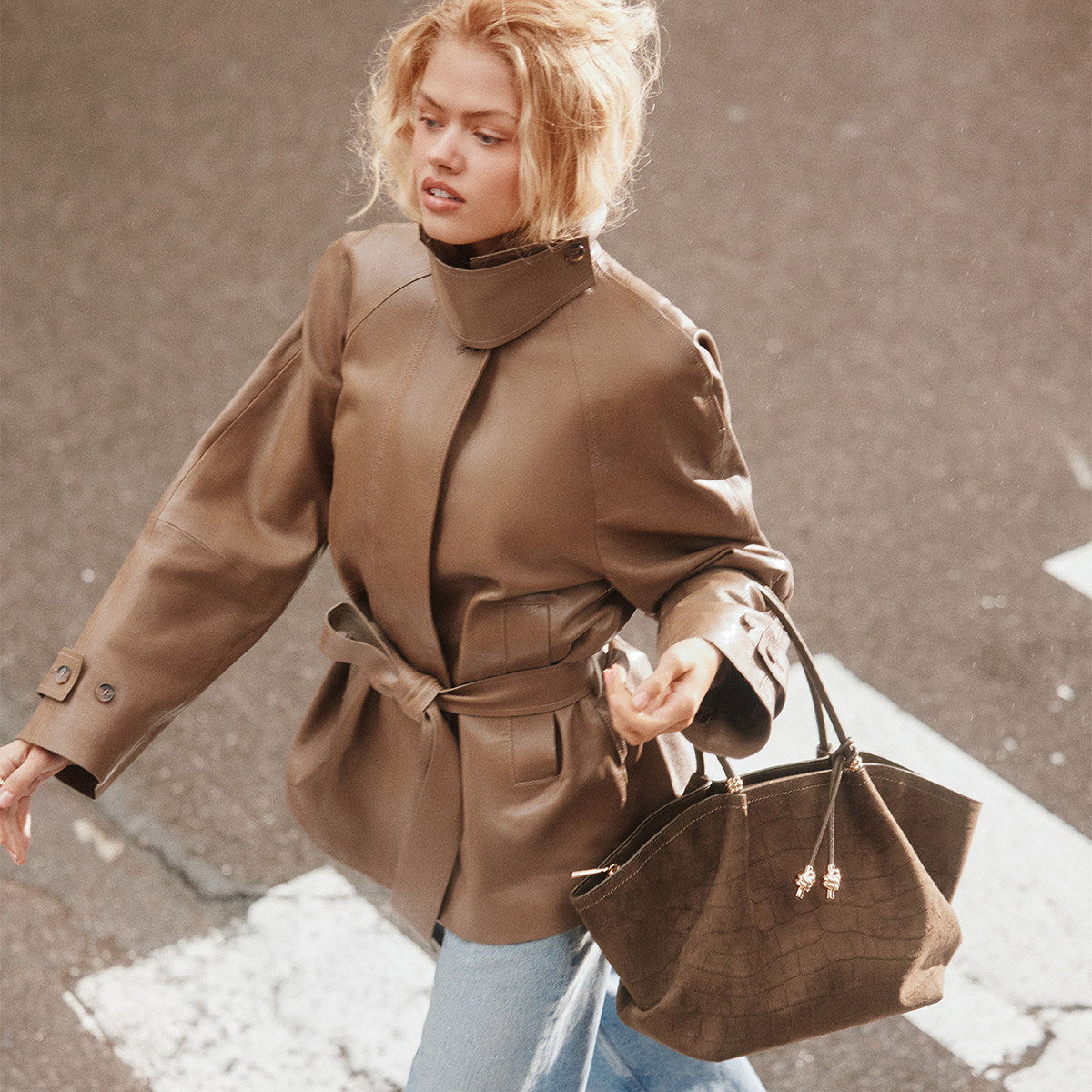 A woman walking outdoors while wearing a structured brown faux-leather belted coat and carrying a matching brown crocodile-embossed tote bag.