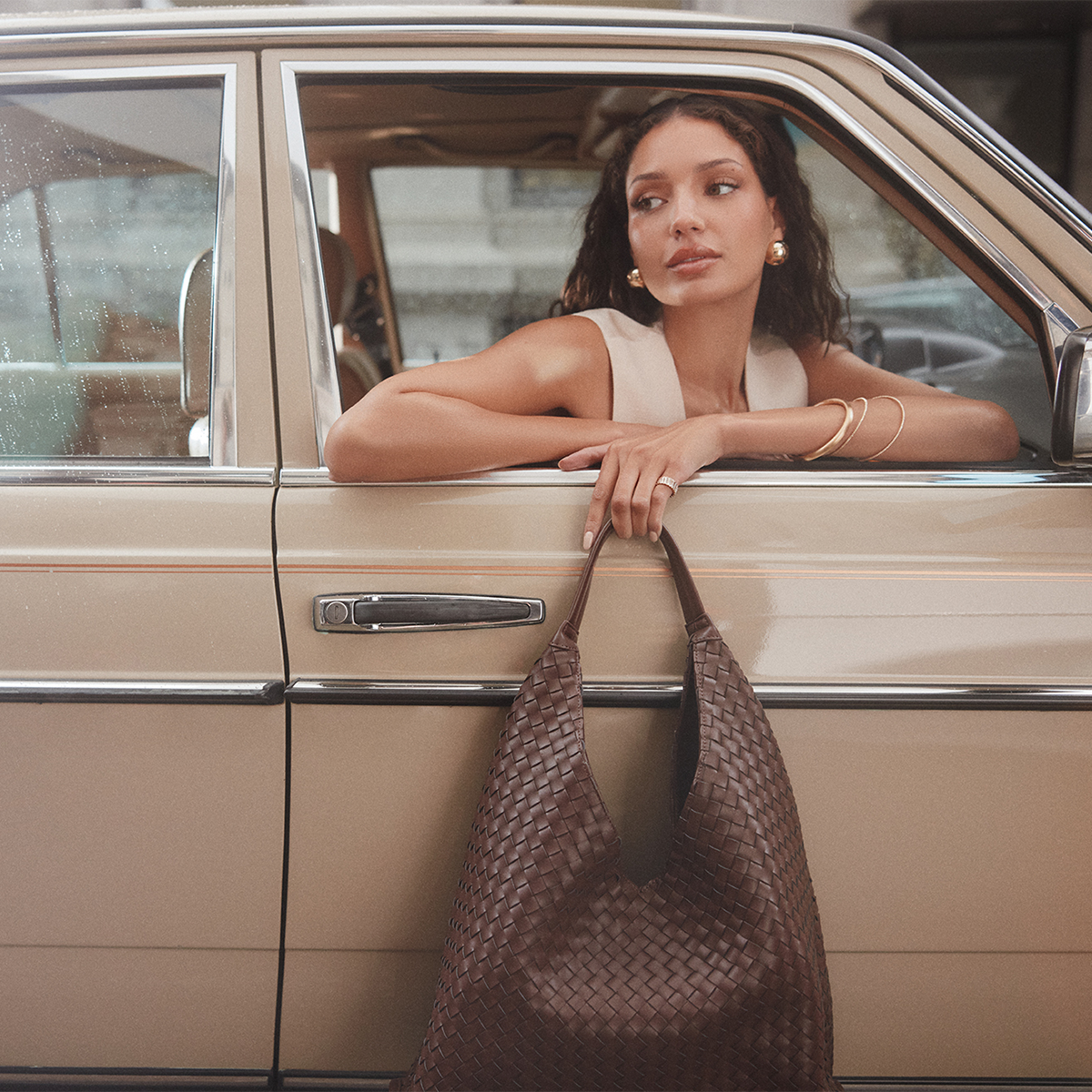 A woman leans out of a beige vintage car window, holding a large woven brown shoulder bag. She is wearing a sleeveless top and gold jewelry.