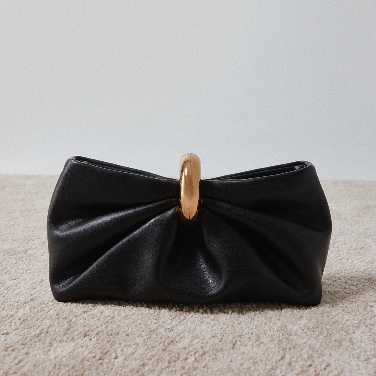 A black leather clutch bag with a gathered, bow-like front and a prominent gold-tone circular handle, resting on a beige carpeted floor against a light gray wall.