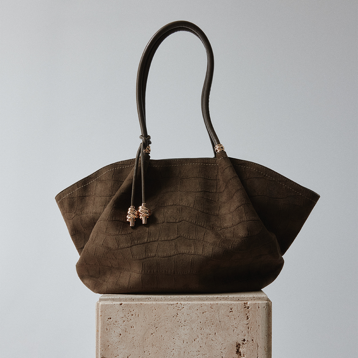 A khaki textured leather handbag with dual top handles and decorative gold-toned bead accents, displayed on a light stone pedestal against a neutral gray background.