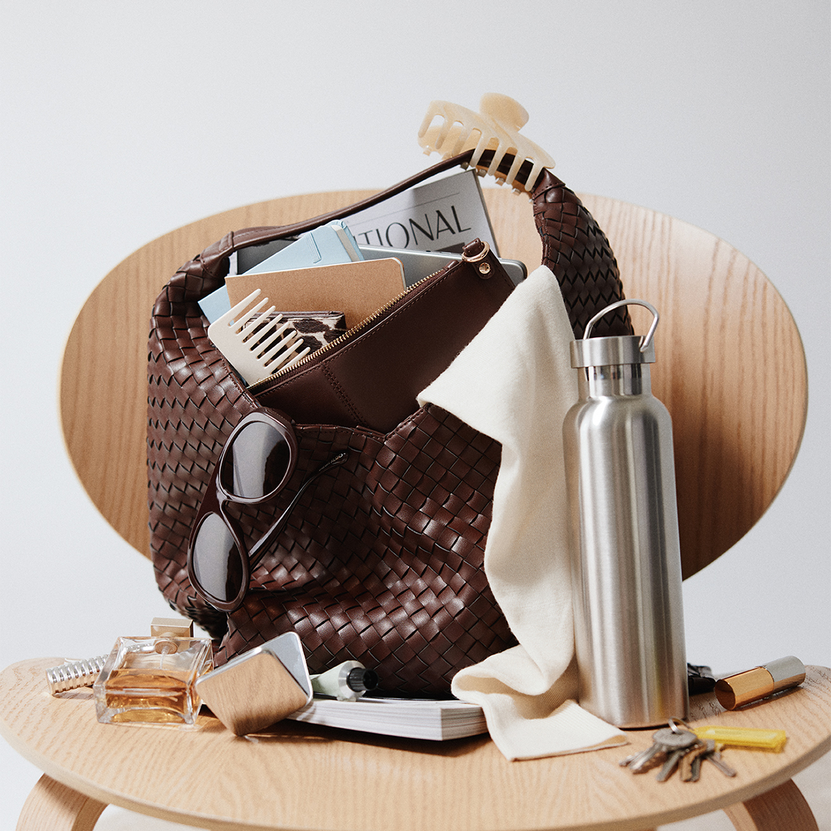 A brown woven handbag sits on a light wood chair, filled with sunglasses, a hair clip, magazines, and a cream cloth. A stainless steel water bottle, keys, and small accessories are arranged around it on the chair against a clean light gray background.