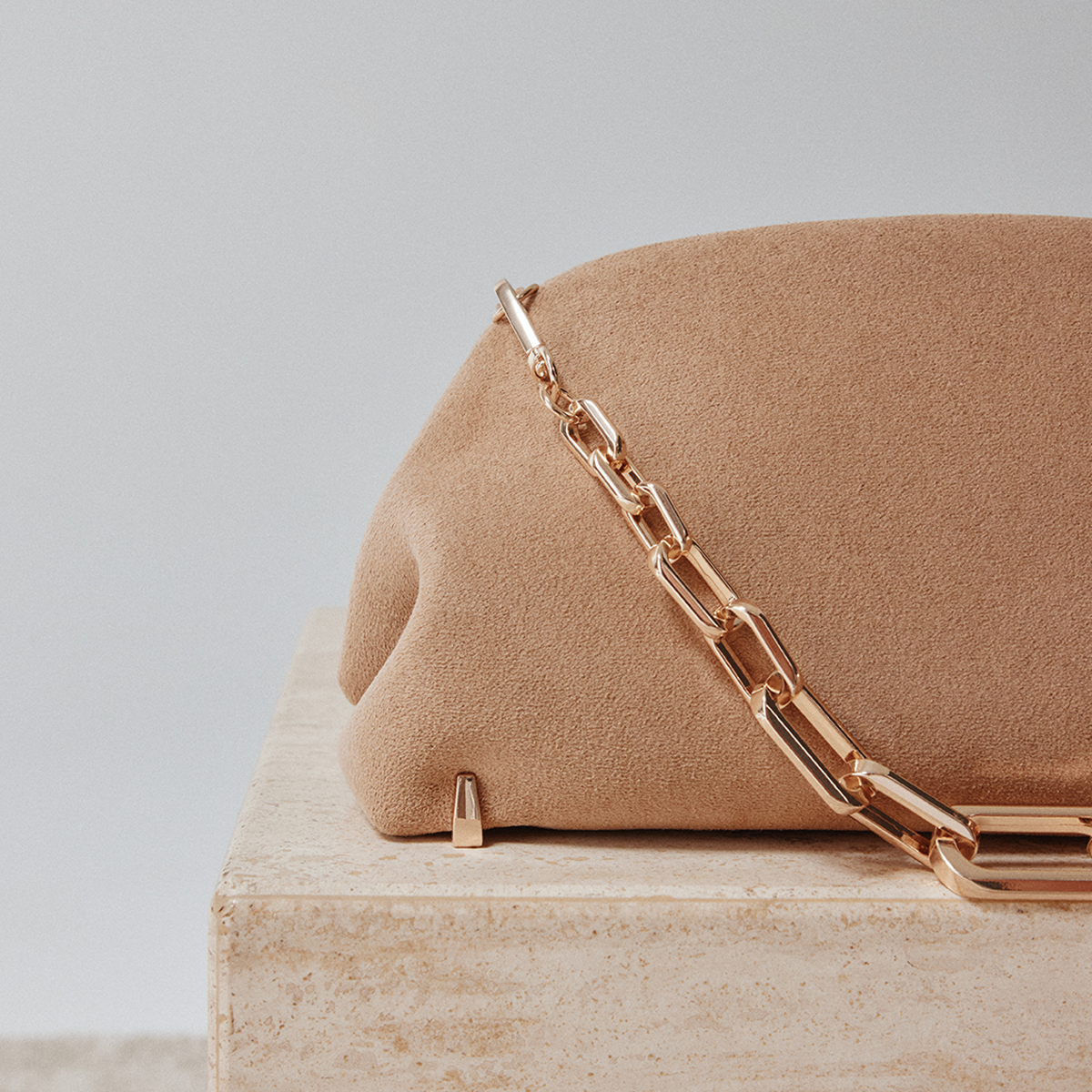 A close-up of a beige suede handbag with a chunky rose gold chain strap, resting on a light beige stone pedestal against a neutral gray background.