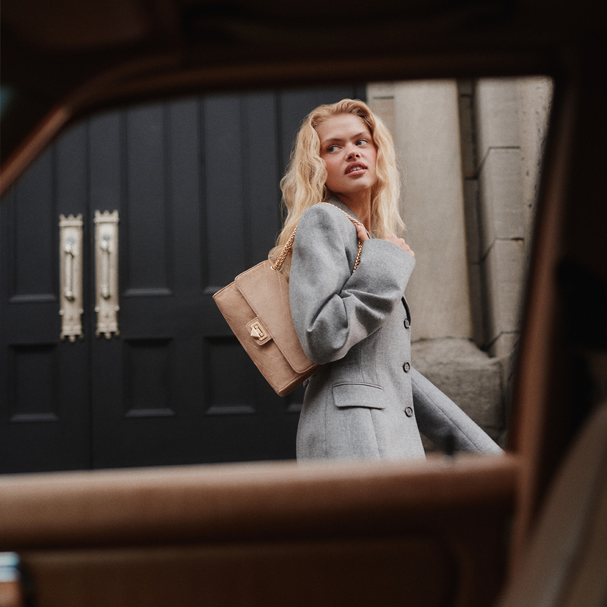 A blonde woman in a gray coat carries a beige shoulder bag with a gold-tone chain strap and turn-lock clasp, photographed through a warm brown frame in front of a dark paneled door.