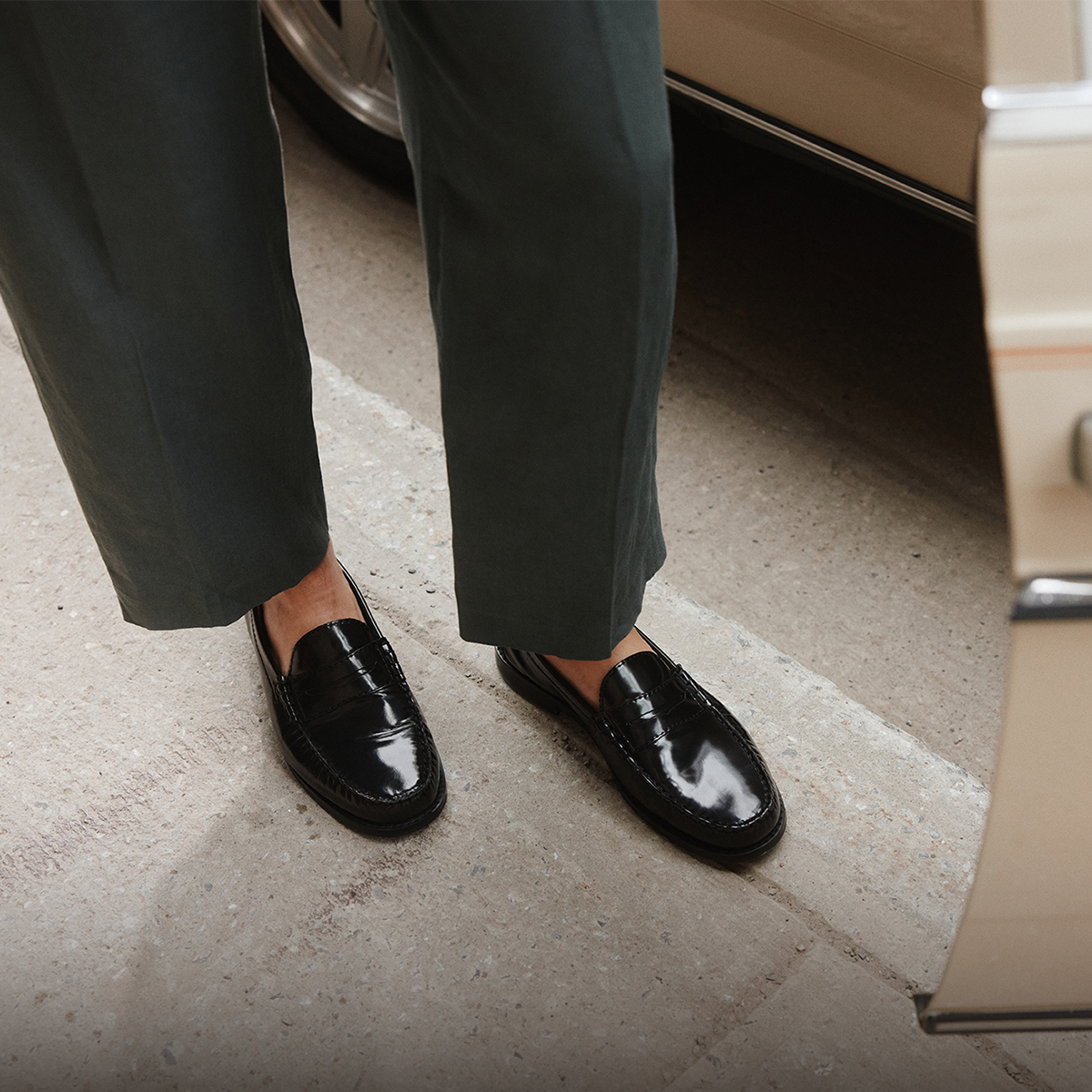 Une personne portant des mocassins penny en cuir verni noir avec un pantalon vert olive foncé, debout sur une surface en béton à côté d’un véhicule beige.