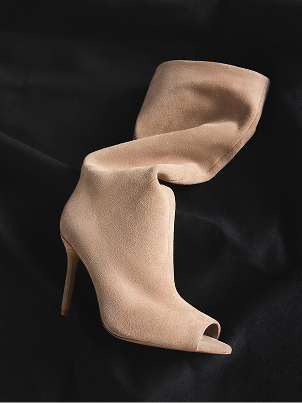 Une botte unique en suède beige nude à bout ouvert et à talon aiguille élevé, drapée sur un tissu noir en arrière-plan.