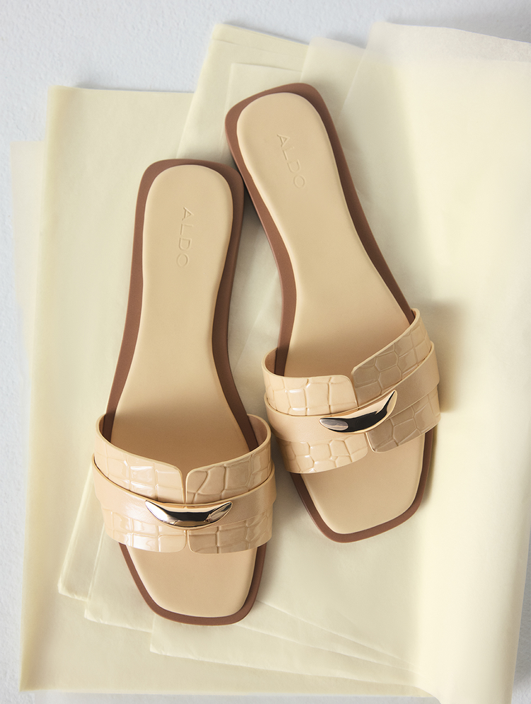 Une paire de sandales de type slide jaunes avec des brides à effet crocodile embossé et un détail métallique poli de couleur or, présentée sur du papier de couleur crème, sur un fond blanc.