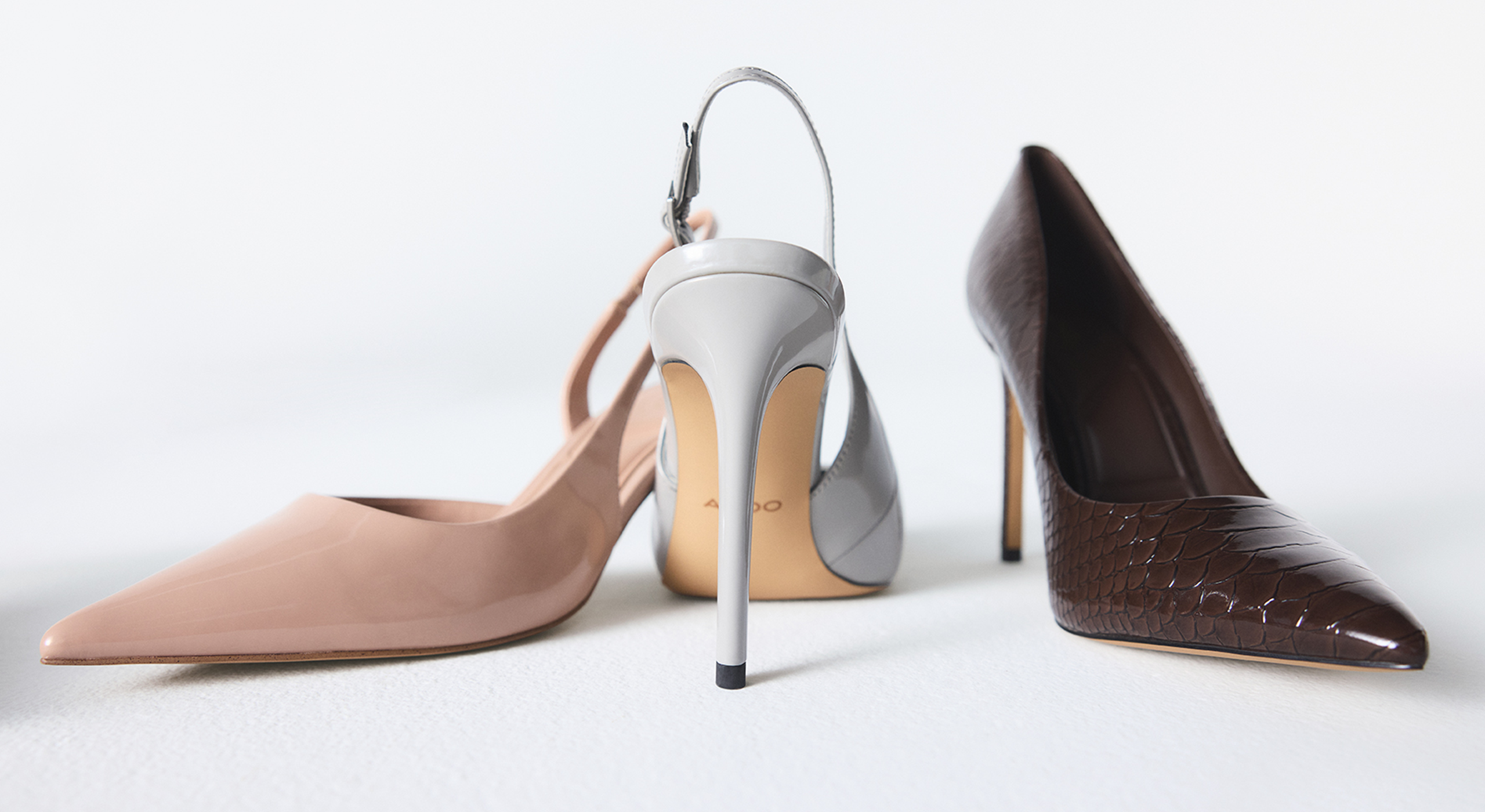 Three elegant pointed-toe high heels arranged on a light gray studio background: a nude beige slingback pump, a light grey slingback pump, and a dark brown croc-embossed pump.