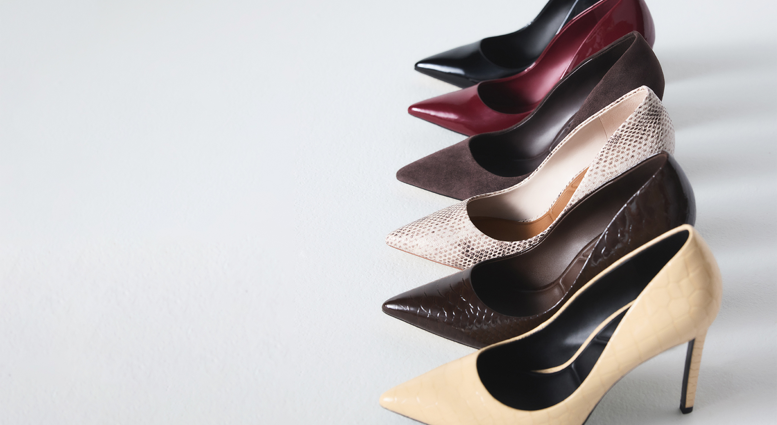 Six pointed-toe high heels in different colors and textures arranged diagonally on a light gray background. The shoes include black, burgundy, dark brown, snakeskin beige, dark brown textured, and yellow.