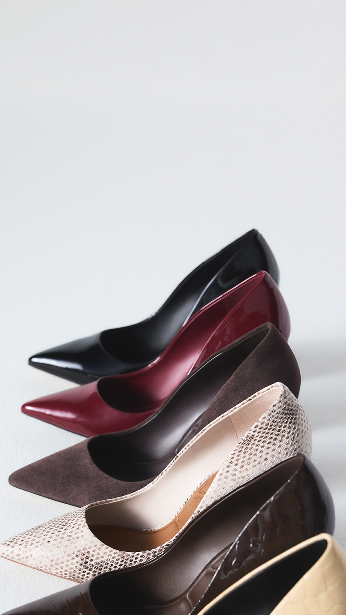 Six pointed-toe high heels in different colors and textures arranged diagonally on a light gray background. The shoes include black, burgundy, dark brown, snakeskin beige, dark brown textured, and yellow.