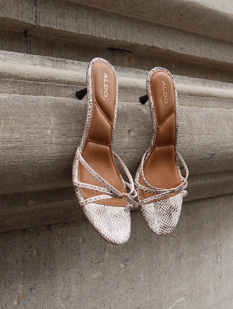 A pair of Aldo brand heeled mule sandals with snakeskin-patterned uppers and tan insoles, resting on a textured concrete ledge.