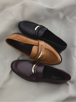 Three penny loafers arranged on draped satin fabric: one black, one tan, and one dark burgundy. Each shoe has a small metallic gold accent on the strap.