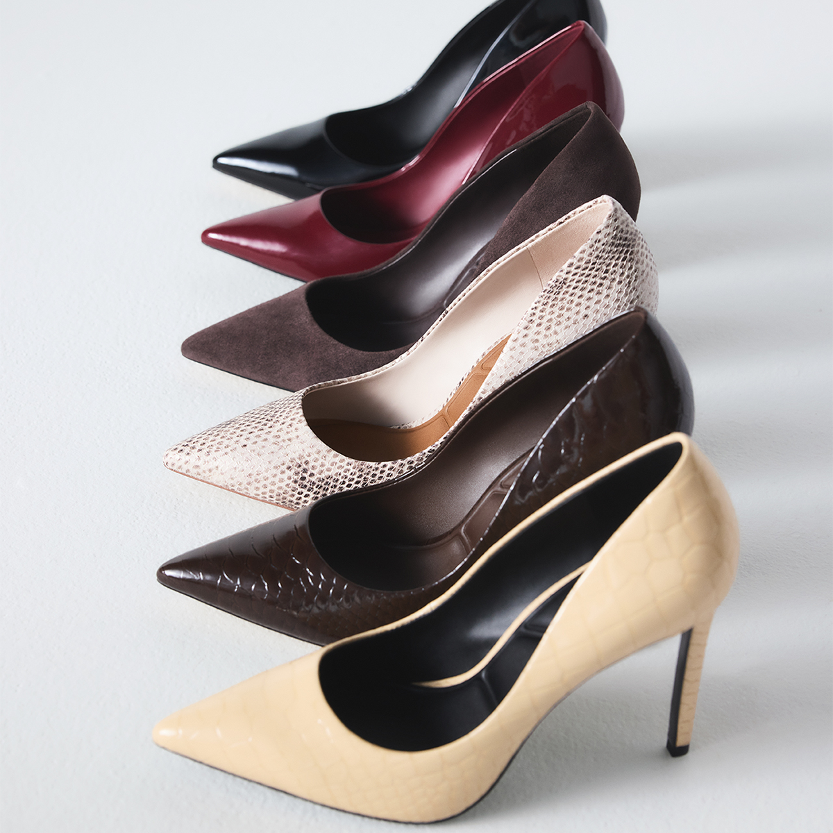 Six pointed-toe high heels arranged diagonally on a light gray background, showing different colors and textures: black patent leather, burgundy patent leather, dark brown suede, snakeskin-pattern beige, dark brown embossed leather, and yellow crocodile-embossed leather.