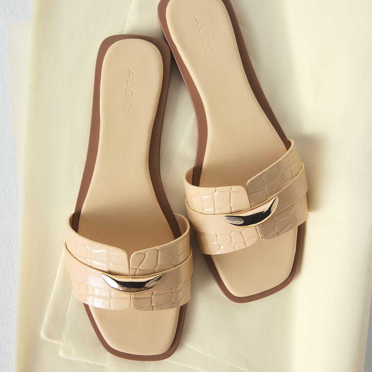A pair of yellow flat slide sandals with crocodile-embossed straps and shiny silver-tone metal accents, displayed on a light cream fabric background. The brand name 'ALDO' is visible on the insoles.