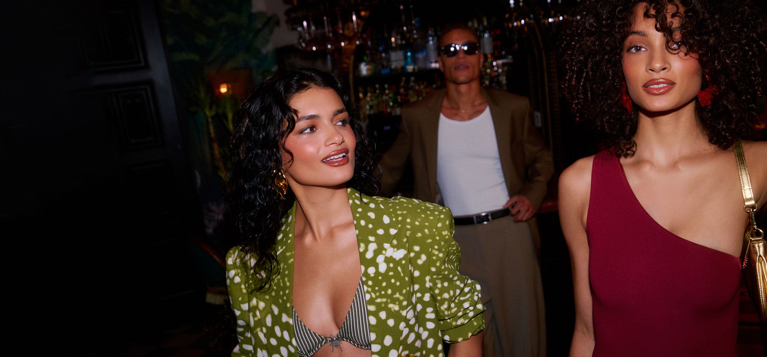 Two women posing in a dimly lit, upscale indoor setting. The woman on the left wears a green and white patterned blazer over a striped bikini top, and the woman on the right wears a deep red one-shoulder dress.