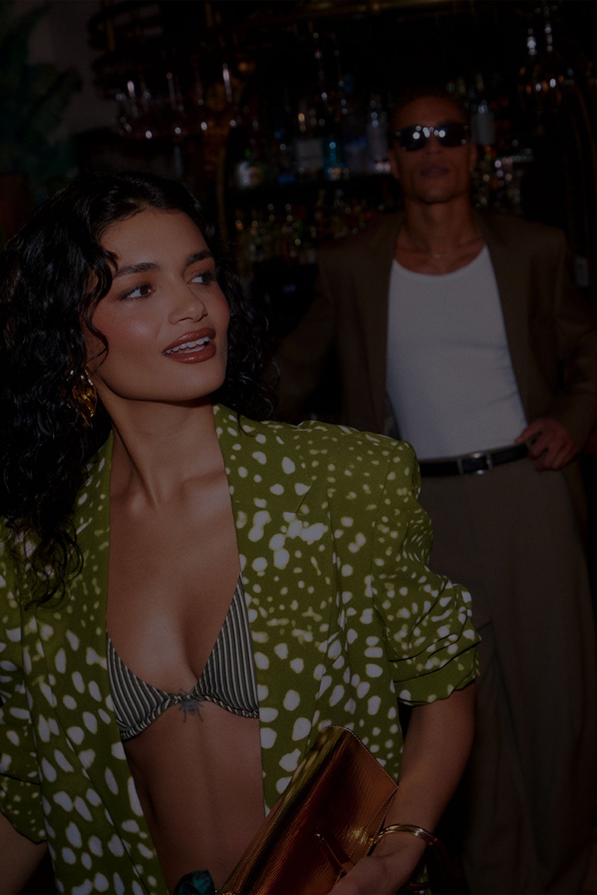 Two women posing in a dimly lit, upscale indoor setting. The woman on the left wears a green and white patterned blazer over a striped bikini top, and the woman on the right wears a deep red one-shoulder dress.