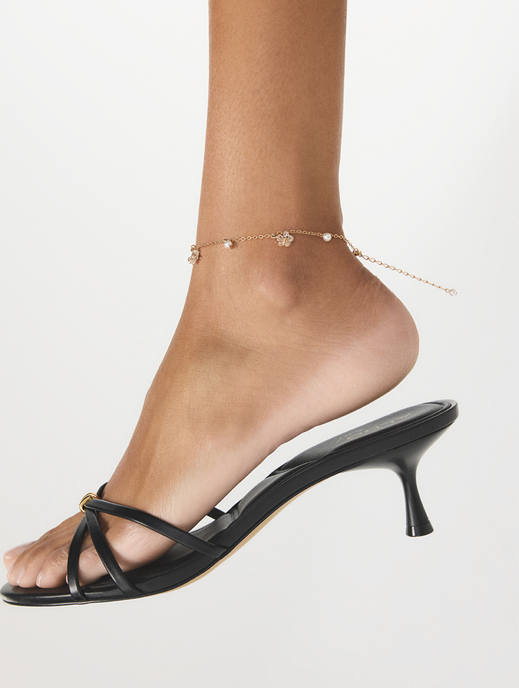 Gros plan du pied d’un mannequin portant un soulier noir à brides et à talon mi-haut, ainsi qu’une fine chaîne de cheville en or rose ornée de petites perles, sur fond blanc.