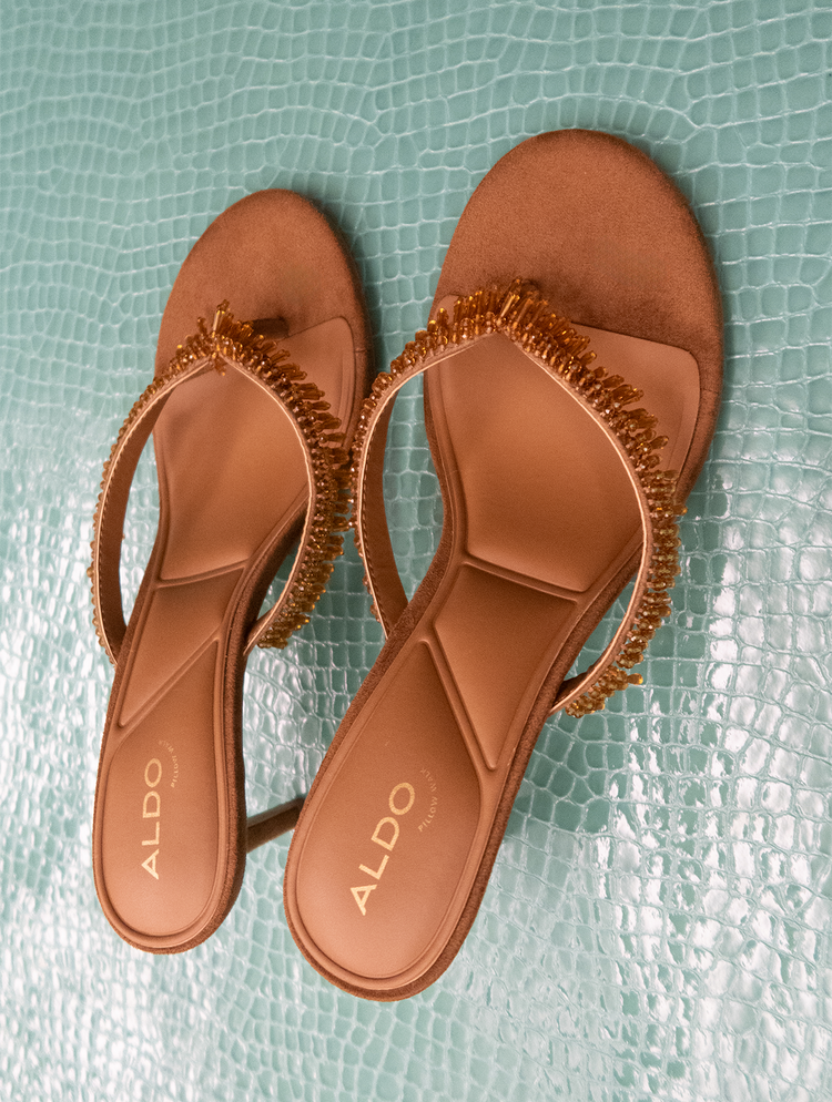 Une paire de tongs plates ALDO en cuir beige, ornées d’une bride perlée, est présentée sur un fond bleu-vert clair à texture effet crocodile.