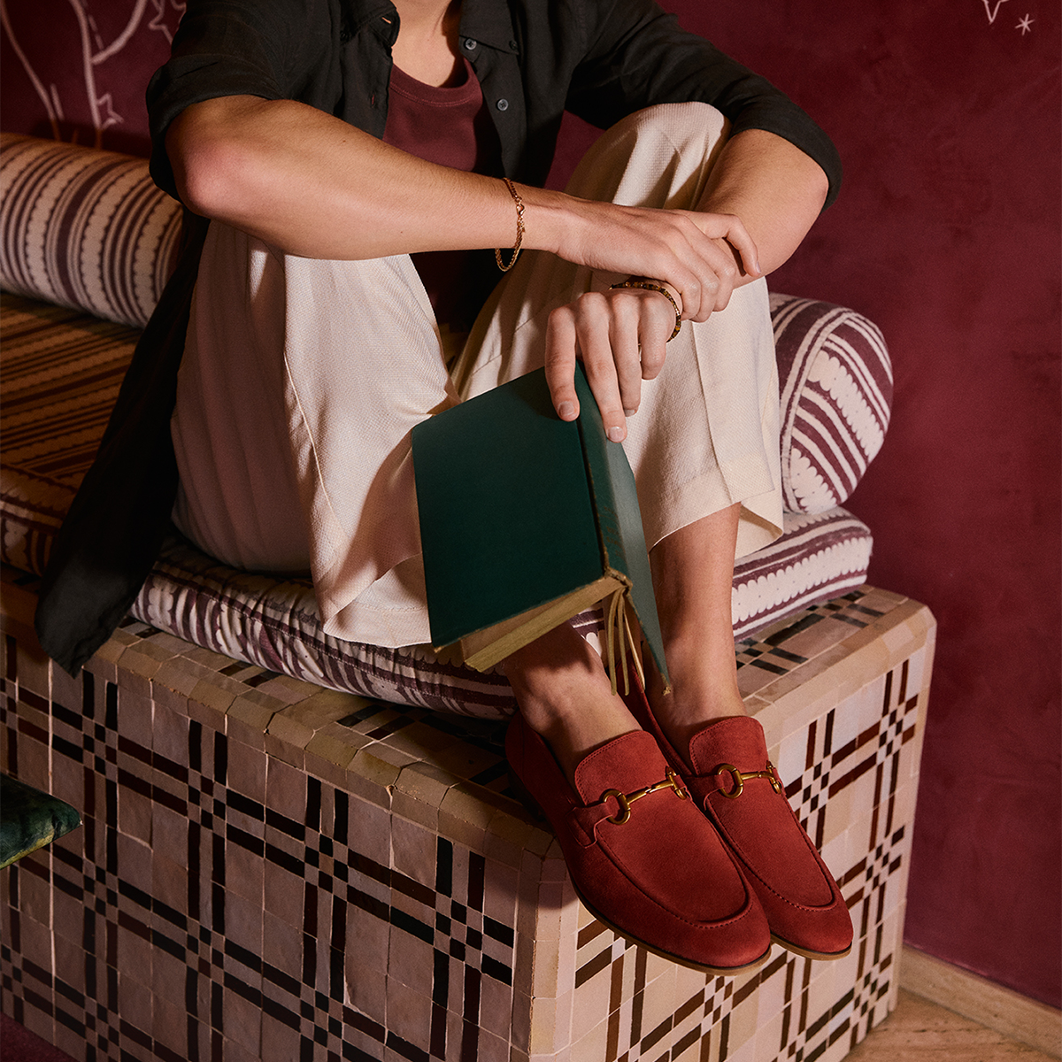 Une personne assise sur une banquette à motifs porte des mocassins en suède rouge ornés d’un mors doré, un pantalon crème et un cardigan noir par-dessus un haut bourgogne. Elle tient un livre vert foncé dans un intérieur à l’ambiance chaleureuse.