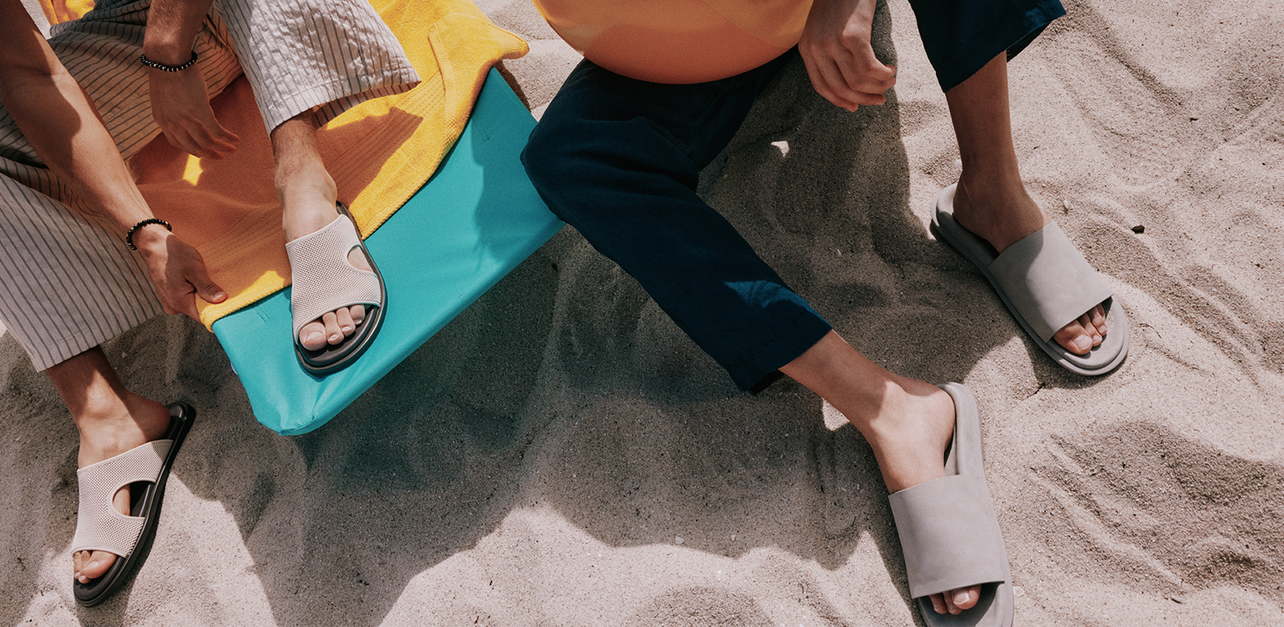 Two men are on the beach. One sits on a blue chair with a yellow towel, wearing striped pants and beige-and-black slide sandals. The other sits on the sand holding a beach ball, wearing navy pants and beige slide sandals.