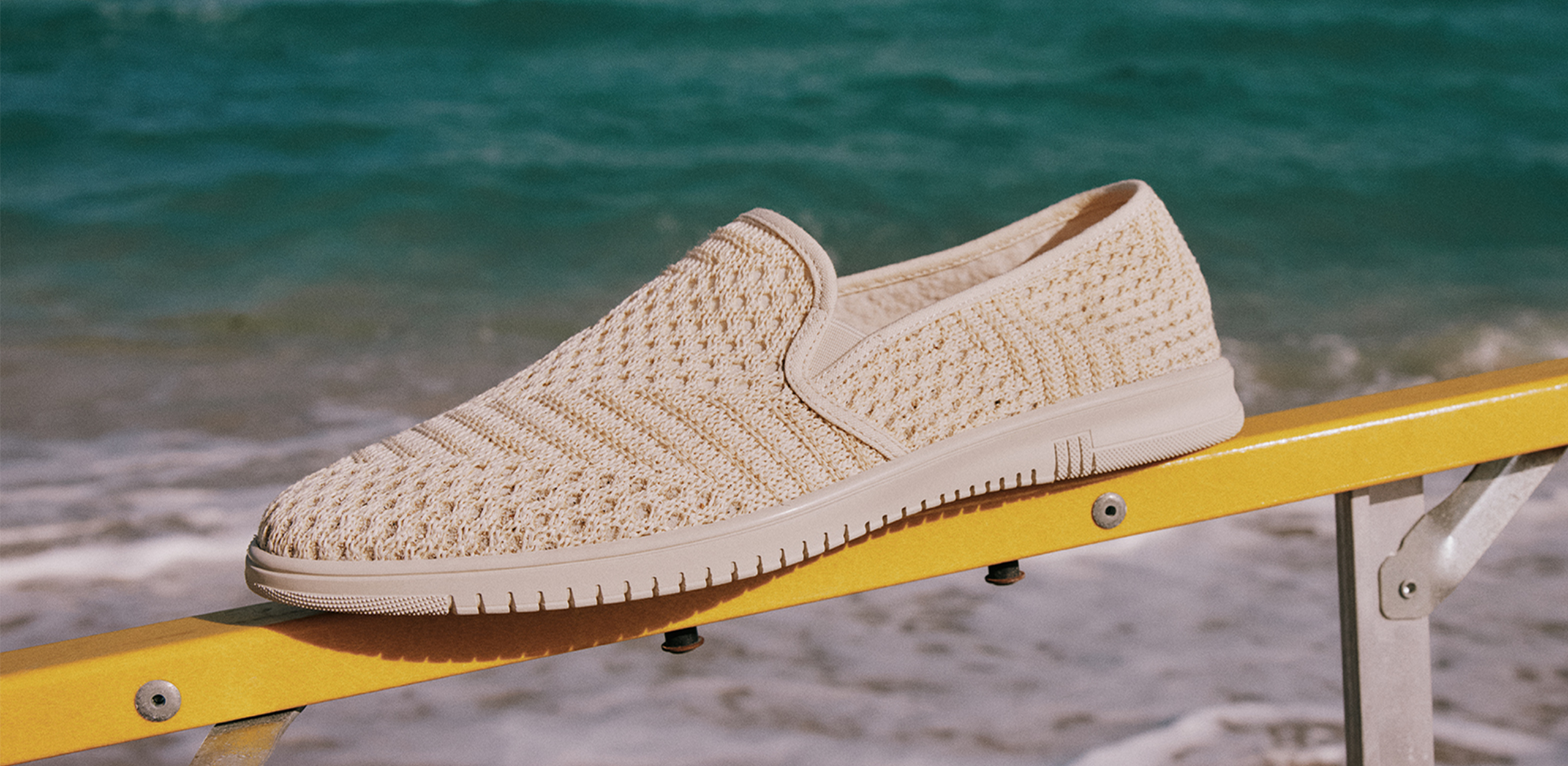 Beige woven shoe on a yellow railing by the ocean.