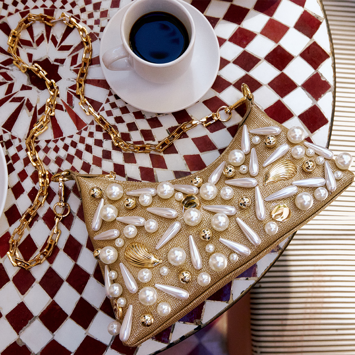Un sac à main décoratif orné de perles et d’accents dorés, posé sur une nappe à carreaux à côté d’une tasse de café.