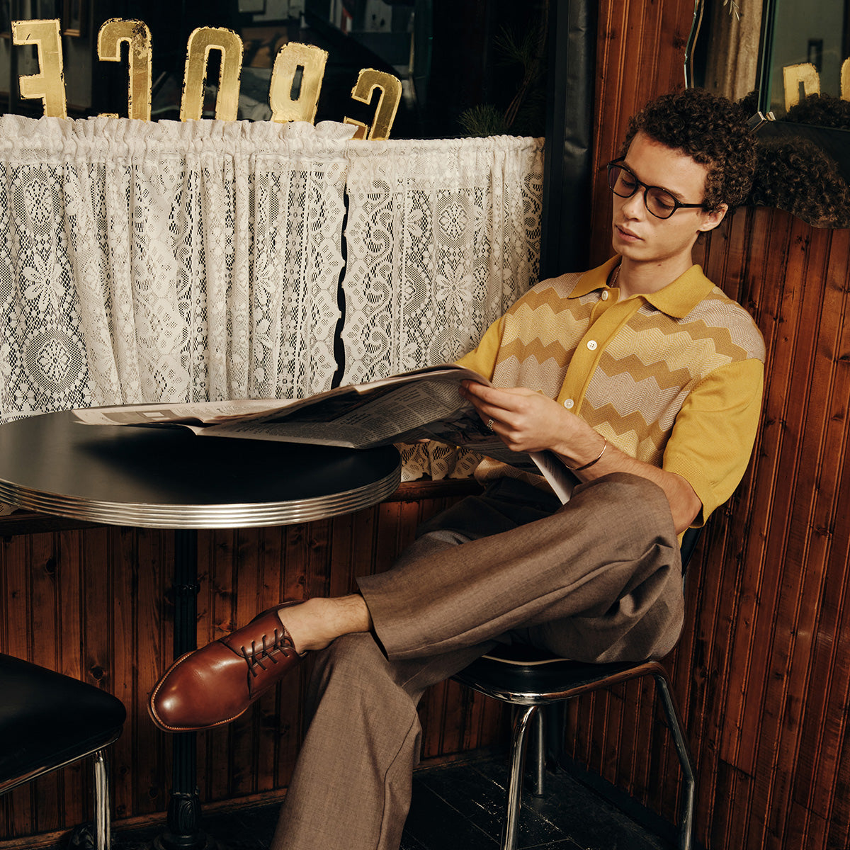 Person sitting wearing brown lace-up dress shoes at a table reading a magazine in a cozy indoor setting.