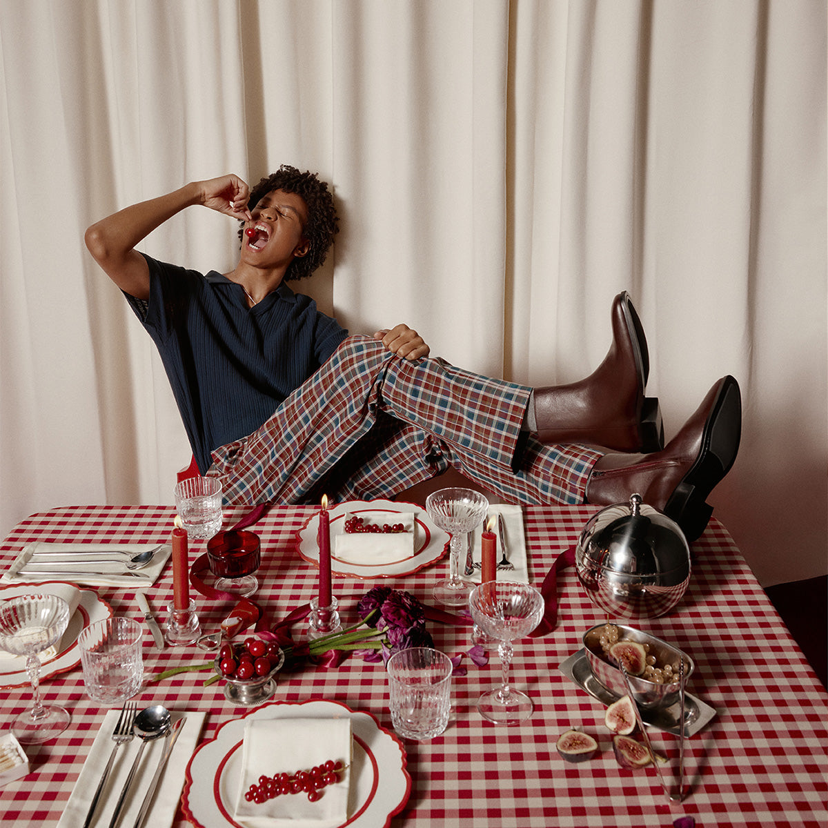 Une personne devant une table recouverte d'une nappe festive, de bougies et de nourriture, portant des bottes bordeaux.