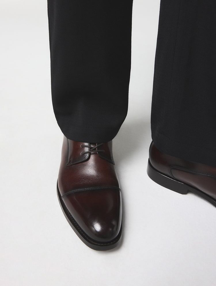 Une vue rapprochée des pieds d’une personne portant des souliers oxford à bout rapporté en cuir brun foncé avec un pantalon habillé noir, photographiés sur un fond gris pâle uni.