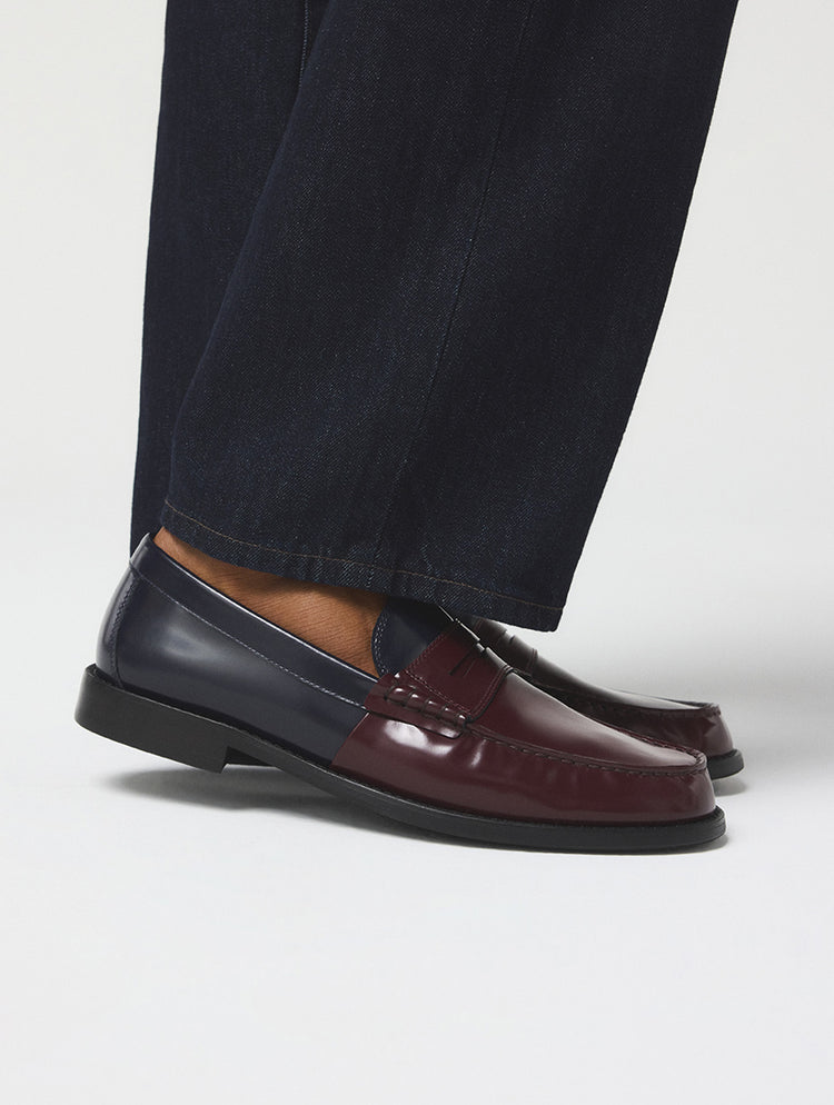 Une paire de mocassins penny pour homme au fini lustré bicolore, combinant des tons bleu marine foncé et bourgogne profond, portée avec un jean bleu foncé sur un fond de studio gris pâle.