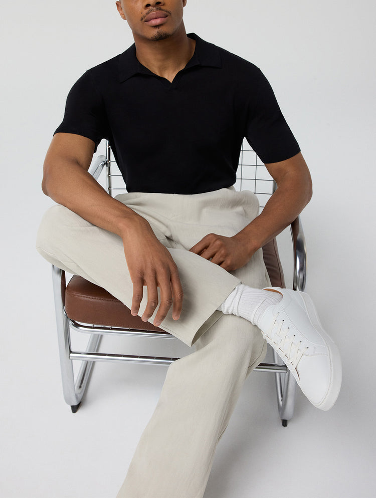 A man seated on a modern chair wearing a black short-sleeve polo shirt, light beige trousers, and white sneakers, photographed against a plain light gray studio background.
