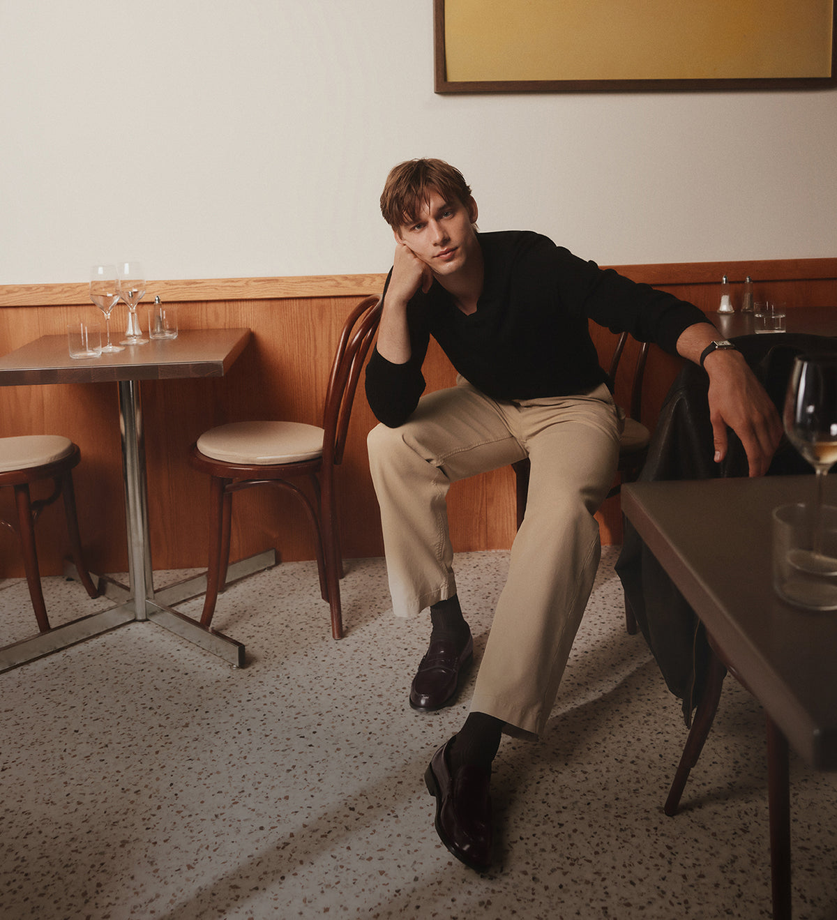 Un homme assis dans un café, portant un haut noir à manches longues, un pantalon beige et des souliers habillés noirs, est appuyé nonchalamment au dossier d’une chaise en bois.