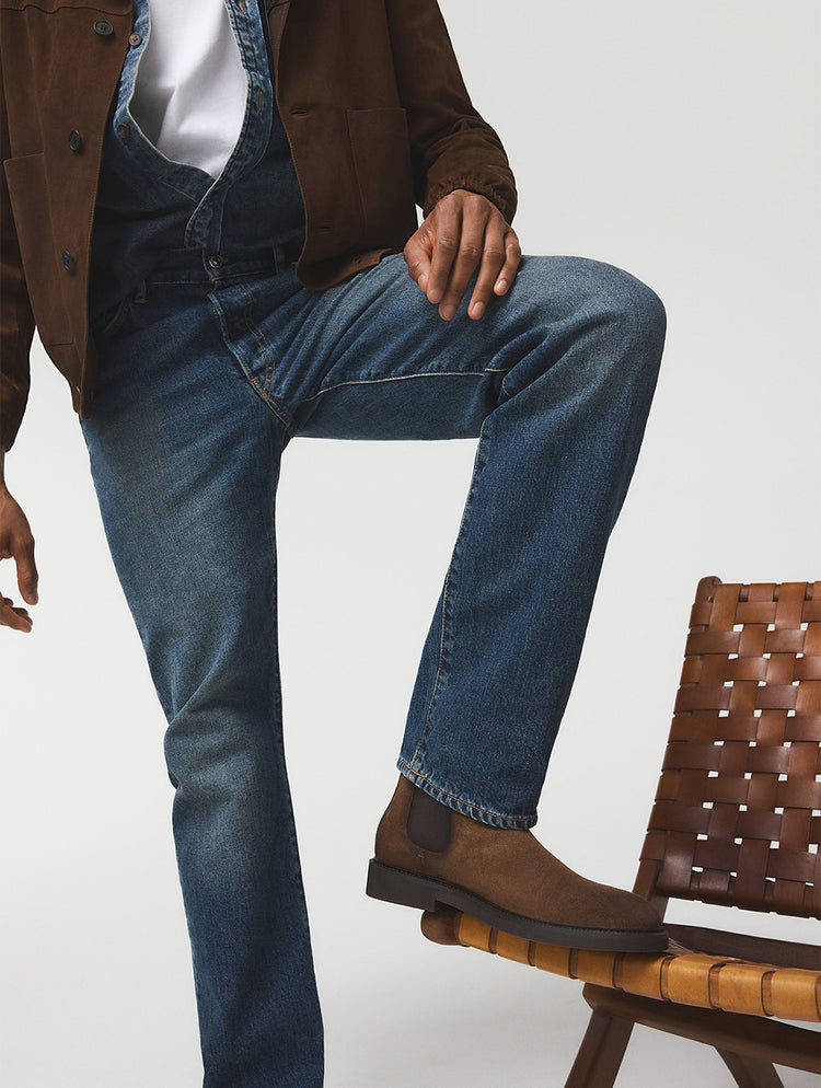 Une personne portant un jean bleu, une veste brune et des bottes en suède brun, assise sur une chaise tressée brune devant un fond de studio gris pâle.