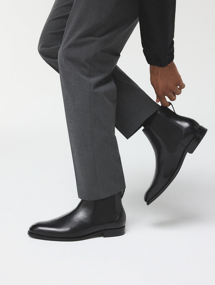 Une vue rapprochée des pieds d’une personne portant un pantalon gris foncé et des bottines Chelsea noires, soulevant l’un des bottillons avec la main afin d’en mettre en valeur le profil latéral, sur un fond de studio blanc épuré.