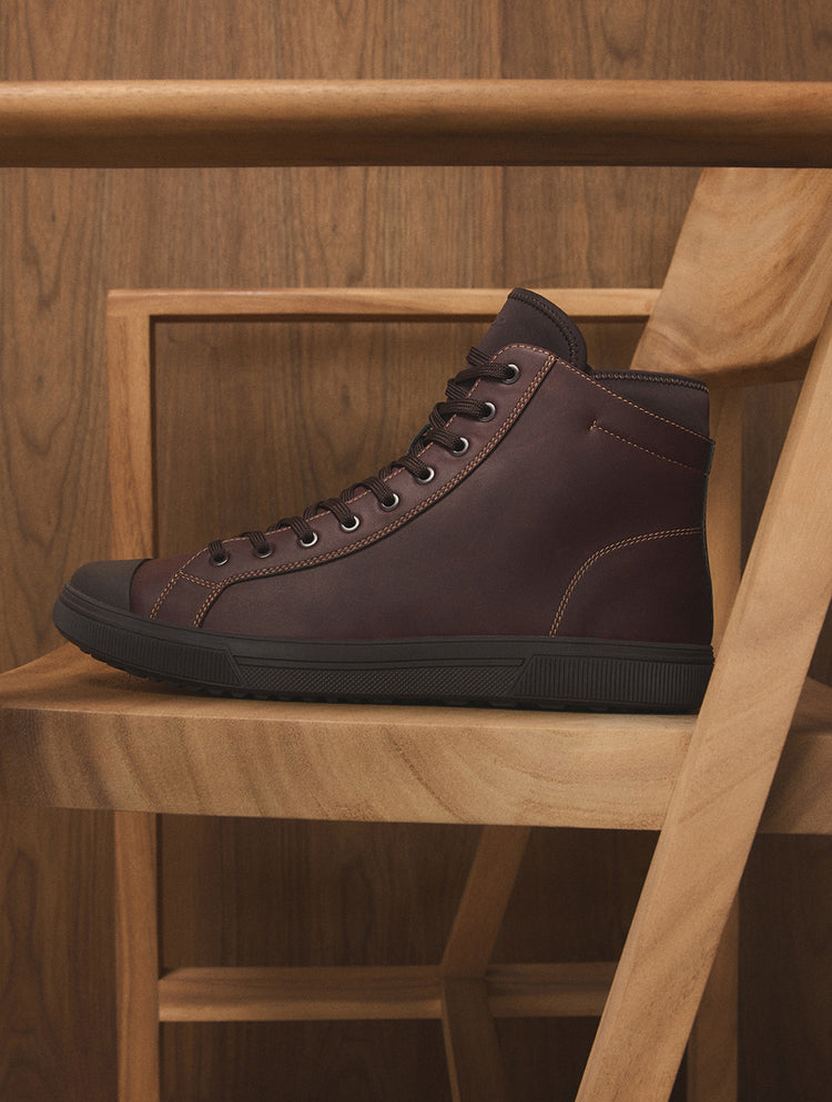 Une espadrille montante en cuir brun foncé dotée de lacets noirs et d’une semelle noire, présentée sur une chaise en bois devant un mur recouvert de panneaux de bois.