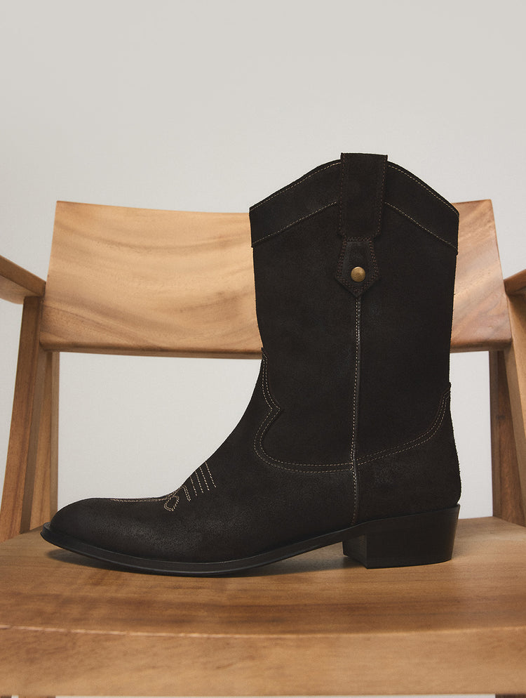 Une botte de style western en suède noir à talon bloc bas et à surpiqûres décoratives, présentée sur une chaise en bois devant un fond gris pâle.