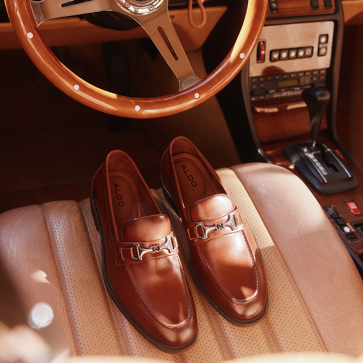 A pair of brown leather Aldo loafers with horsebit straps, displayed on a tan leather car seat with a vintage wooden steering wheel and dashboard in the background.